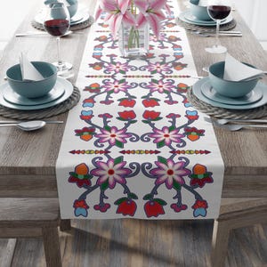 May include: White table runner with a colorful floral pattern. The pattern features pink, red, orange, yellow, blue, and green flowers and leaves. The runner is on a wooden table with place settings.