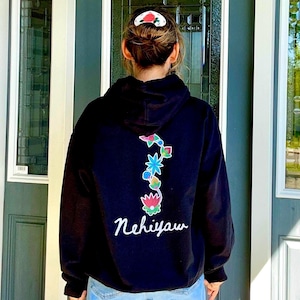 May include: Black hoodie featuring a vibrant floral design on the back, with the word "Nehiyaw" in white script. The floral pattern incorporates pink, blue, and green hues. The person is wearing a white and red hair clip.