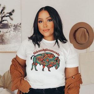 Peut inclure: T-shirt blanc avec un motif de bison coloré orné de motifs floraux rouges, turquoise et verts. Le texte "ahkamêgîhtamowin" est au-dessus du bison. Vêtement décontracté.