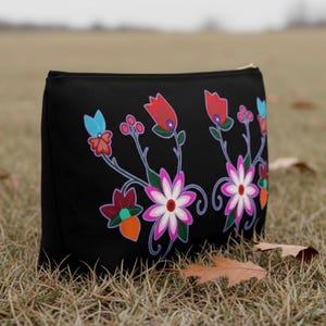 May include: Black zippered pouch featuring a colorful floral design. The flowers are in shades of red, pink, orange, and blue, with green stems and leaves. The pouch is sitting on a bed of dry grass.