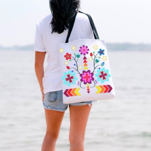 May include: A white tote bag with black straps features a colorful floral design with red, pink, blue, yellow, and green flowers. The flowers are arranged in a symmetrical pattern with arrows at the bottom.