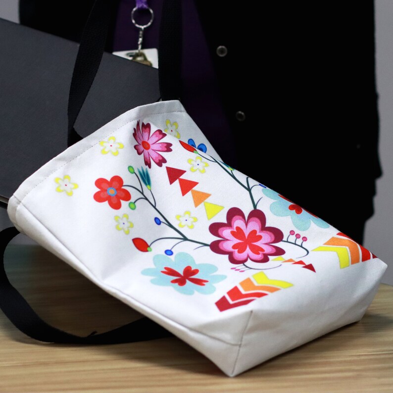 May include: White tote bag with a colorful floral design featuring red, pink, yellow, blue, and green flowers. The bag has a black handle and is made of canvas.