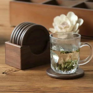 Peut inclure: Un ensemble de six dessous de verre en bois brun foncé dans un support en bois assorti. Une tasse en verre transparent avec une poignée repose sur l'un des dessous de verre, remplie d'eau et d'herbes vertes. Une fleur blanche est en arrière-plan.