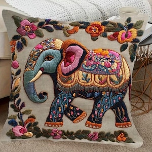 Peut inclure: Un coussin carré en tissu beige clair orné d'une broderie complexe représentant un éléphant central dans des tons de bleu, rose et doré. L'éléphant est entouré d'une bordure décorative composée de vignes et de feuilles florales colorées.