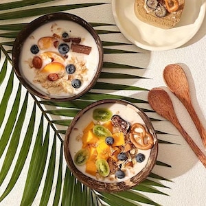 Peut inclure: Deux bols en coque de noix de coco brun foncé remplis de yaourt blanc, garnis de myrtilles, de tranches de pêche, de morceaux de mangue, de raisins verts, d'amandes, de morceaux de chocolat et d'un petit bretzel. Deux cuillères en bois.