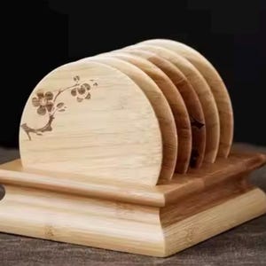 Peut inclure: Un ensemble de six dessous de verre en bois dans un support. Les dessous de verre sont brun clair et semi-circulaires, avec un motif de branche florale gravé sur l'un d'eux. Le support est une base rectangulaire marron foncé avec un design incurvé.