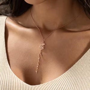 Peut inclure: Un collier couleur or rose composé d'une chaîne fine et d'un pendentif vertical. Le pendentif présente un petit motif floral en haut, suivi d'un nom écrit en lettres cursives qui se prolonge vers le bas dans un design linéaire.