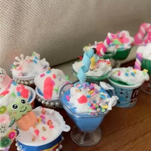 Könnte beinhalten: Eine Sammlung von Miniatur-Eisbechern in verschiedenen Farben und Designs. Die Eisbecher sind mit Streuseln, Süßigkeiten und verschiedenen Toppings verziert. Einige der Eisbecher befinden sich in klaren Glasschalen, während andere in Papierbechern sind.