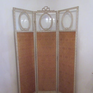 May include: A three-panel room divider with a white painted finish. Each panel features a carved oval frame with a clear glass insert. The panels are made of wood and have a light brown fabric backing.