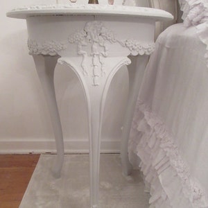 May include: A white, ornate side table with a round top and three curved legs. The table features detailed floral and scrollwork carvings. A brass candelabra with pink crystal accents sits on top of the table. The table is next to a bed with ruffled bedding.
