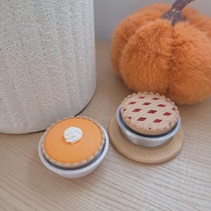 May include: Two miniature toy pies in small bowls. One pie is orange with a white dollop of whipped cream on top. The other pie has a tan lattice crust revealing a red filling underneath. Both pies are set next to a fuzzy orange pumpkin.