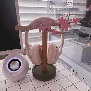 May include: A decorative headphone stand in a cherry blossom design, holding a pair of light pink headphones. The stand is made of wood and features pink flowers. A white speaker and a pink mouse are also visible.