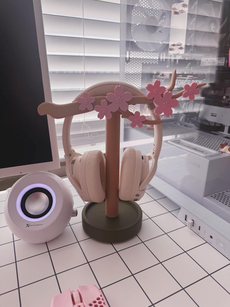 May include: A decorative headphone stand in a cherry blossom design, holding a pair of light pink headphones. The stand is made of wood and features pink flowers. A white speaker and a pink mouse are also visible.