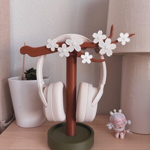 May include: A decorative headphone stand with a brown stem and a green base. The stand features a branch with white flower accents. White headphones are displayed on the stand. A small figurine is on the wooden surface.