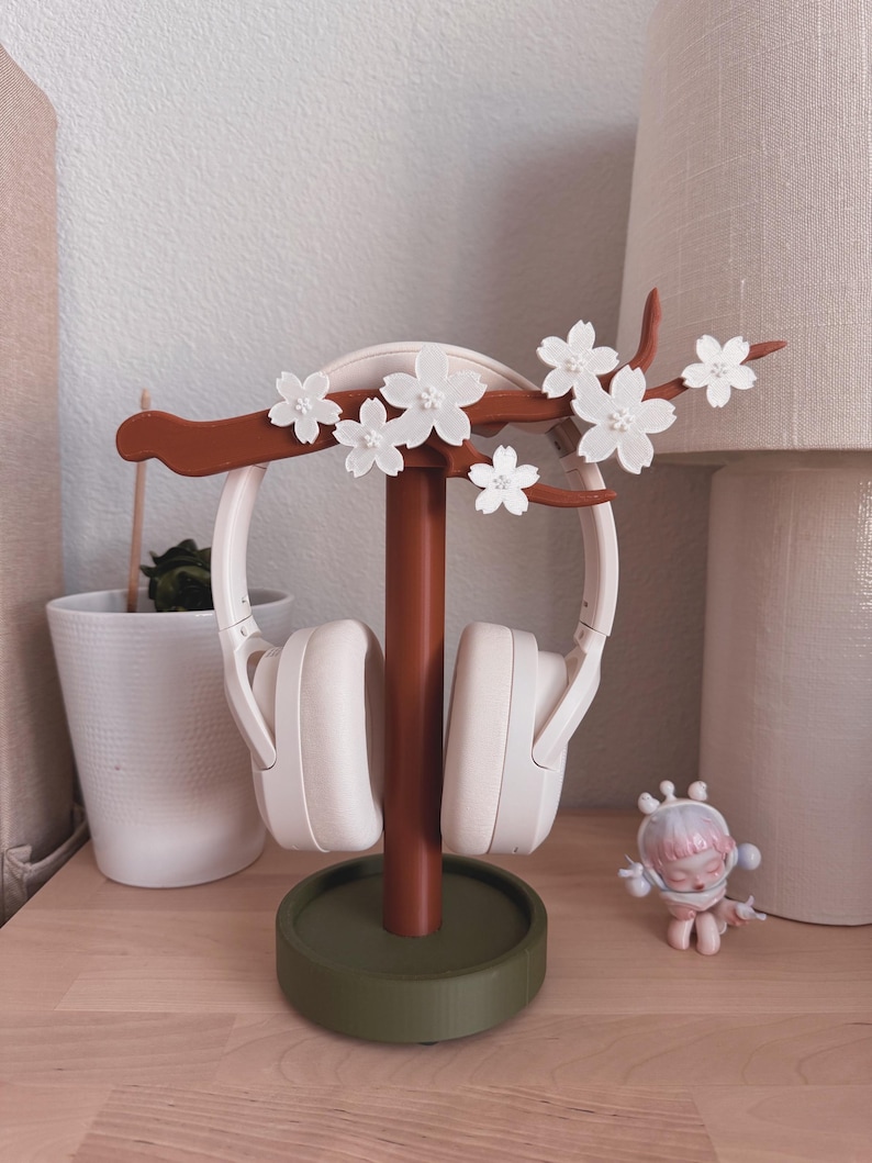 May include: A decorative headphone stand with a brown stem and a green base. The stand features a branch with white flower accents. White headphones are displayed on the stand. A small figurine is on the wooden surface.