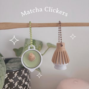 May include: Two Matcha Clickers hanging from a wooden rod. One is light green with a beige center, the other is beige with a white base. Both are attached to a chain. The text "Matcha Clickers" is above.