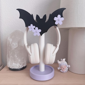 May include: A black bat-shaped headphone stand with lavender flower details. The stand holds white headphones and sits on a lavender base. A small figurine is next to the stand.