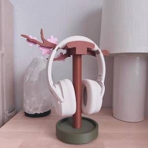 May include: A white headset rests on a decorative headphone stand. The stand is brown with a green base and features a branch with pink flower accents. The stand is on a light wood surface.
