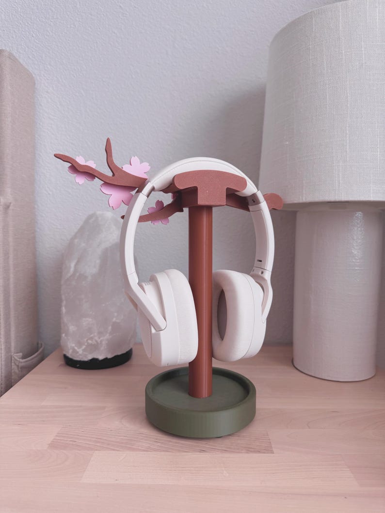 May include: A white headset rests on a decorative headphone stand. The stand is brown with a green base and features a branch with pink flower accents. The stand is on a light wood surface.
