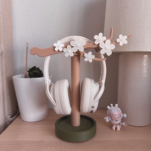 May include: A decorative headphone stand in a soft color palette. The stand features a brown branch with white flower accents, a central column, and a green circular base. White headphones are displayed on the stand.