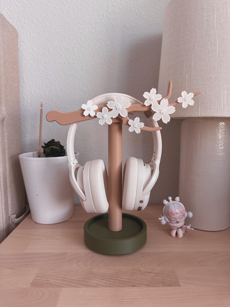 May include: A decorative headphone stand in a soft color palette. The stand features a brown branch with white flower accents, a central column, and a green circular base. White headphones are displayed on the stand.