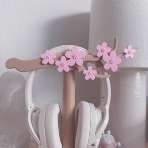 May include: A white headset rests on a pink and brown headphone stand. The stand is decorated with pink cherry blossom flowers. The stand is on a green circular base. A lamp and a small figurine are in the background.