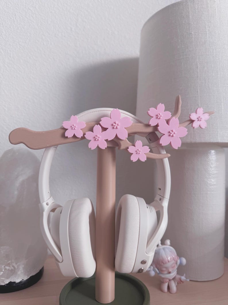 May include: A white headset rests on a pink and brown headphone stand. The stand is decorated with pink cherry blossom flowers. The stand is on a green circular base. A lamp and a small figurine are in the background.