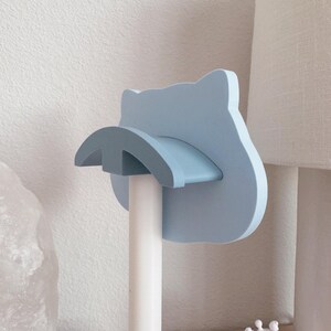 Op de afbeelding: Een lichtblauwe katvormige toiletpapierhouder. De houder heeft een gebogen plank en een ronde basis. Hij is gemonteerd op een witte paal. De achtergrond omvat een witte muur en een lamp.