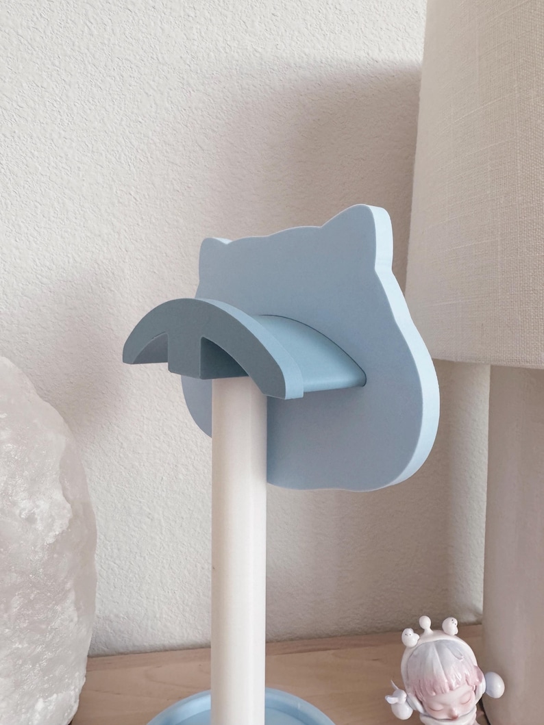 Op de afbeelding: Een lichtblauwe katvormige toiletpapierhouder. De houder heeft een gebogen plank en een ronde basis. Hij is gemonteerd op een witte paal. De achtergrond omvat een witte muur en een lamp.
