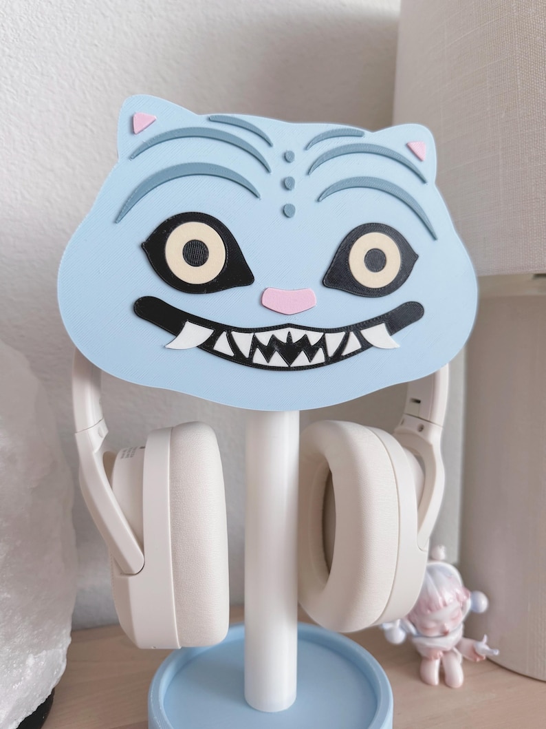 Op de afbeelding: Een lichtblauwe katvormige hoofdtelefoonstandaard met een witte basis en een lichtblauwe cirkelvormige basis. De kattenkop heeft zwarte en beige ogen, een roze neus en een witte grijns. Witte hoofdtelefoon hangt aan de standaard.