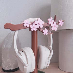 May include: A decorative headphone stand featuring a brown branch adorned with pink cherry blossoms. The stand holds a pair of white headphones. The base is a muted green color, providing a stylish and functional accessory for any desk or bedside table.