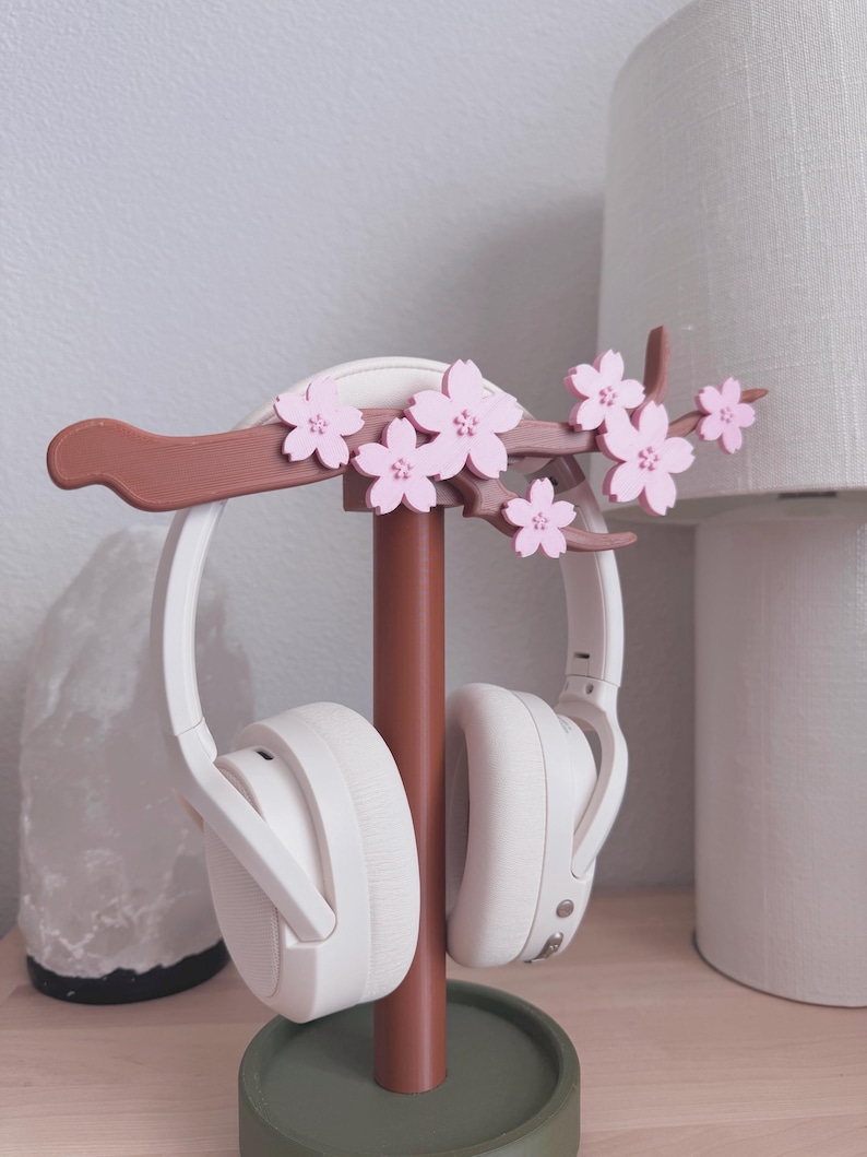 May include: A decorative headphone stand featuring a brown branch adorned with pink cherry blossoms. The stand holds a pair of white headphones. The base is a muted green color, providing a stylish and functional accessory for any desk or bedside table.