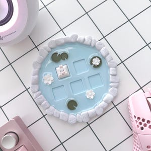 May include: A light blue, pond-shaped keyboard keycap holder with white stone-like edging, lily pads, and flowers. A keycap with a bunny design is in the center. A pink keyboard, mouse, and speaker are also visible.