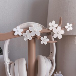 May include: A white headset rests on a decorative headphone stand. The stand is light brown and features a branch design with white flower accents. The stand is on a wooden surface.