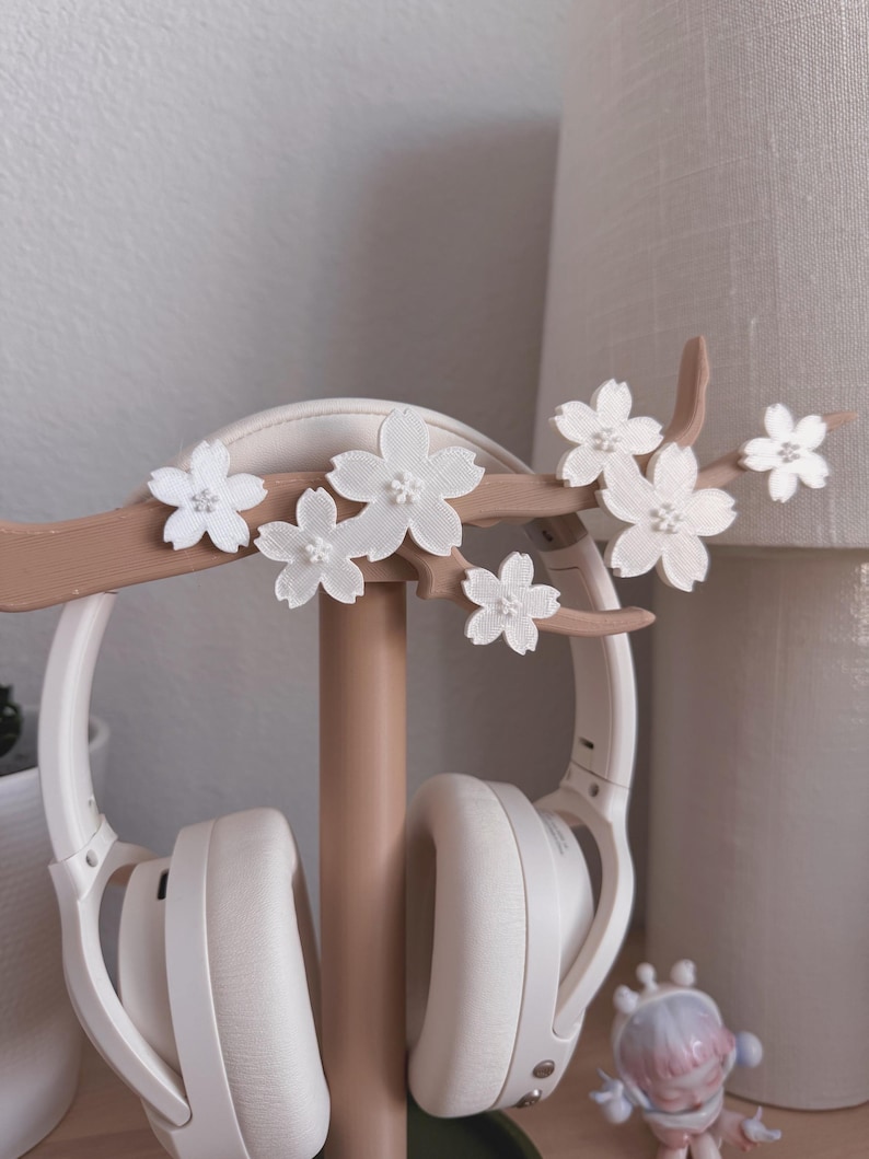 May include: A white headset rests on a decorative headphone stand. The stand is light brown and features a branch design with white flower accents. The stand is on a wooden surface.