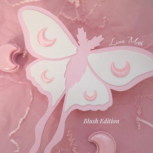 May include: A layered paper art piece featuring a large pink and white moth with crescent moon accents. The moth is set against a pink background with the text "Luna Moth" and "Blush Edition".