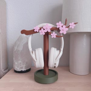 May include: A decorative headphone stand featuring a brown stem and a green base, adorned with pink cherry blossom accents. White headphones are displayed on the stand. The stand is on a light wood surface.