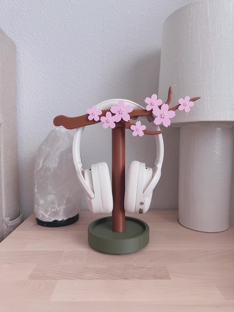 May include: A decorative headphone stand featuring a brown stem and a green base, adorned with pink cherry blossom accents. White headphones are displayed on the stand. The stand is on a light wood surface.