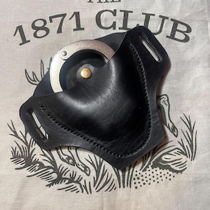 May include: Black leather handcuff holder with a silver handcuff inside. The holder has a stitched design and a brass rivet. The background is a tan cloth with the text "THE 1871 CLUB" printed in black.