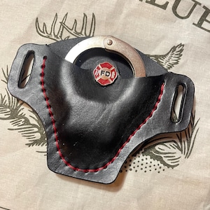May include: Black leather handcuff holder with red stitching and a silver handcuff. The holder has a red and white FD badge. The background has the text "THE 1871 CLUB" and a deer antler graphic.