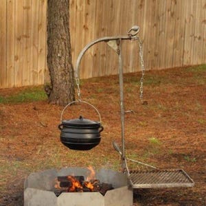 May include: A black cast iron pot with a lid hangs from a metal arm over a hexagonal fire pit. The setup includes a metal chain for height adjustment and a rectangular metal grate attached to the side for cooking over an open fire.