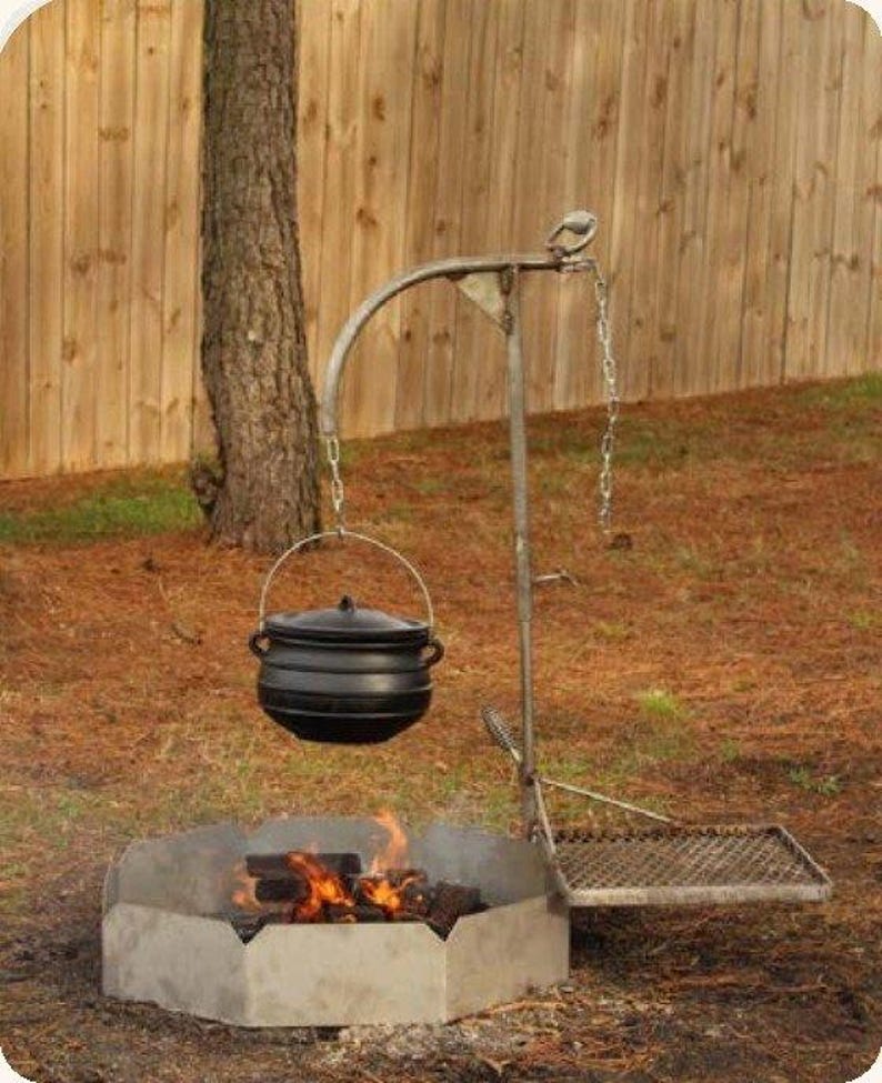 May include: A black cast iron pot with a lid hangs from a metal arm over a hexagonal fire pit. The setup includes a metal chain for height adjustment and a rectangular metal grate attached to the side for cooking over an open fire.