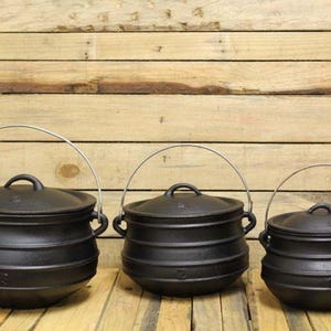 May include: Three black cast iron pots of varying sizes, each featuring a rounded body, a matching lid with a small handle, and a silver-coloured metal wire bail handle attached to two side lugs. The pots are arranged in a row.