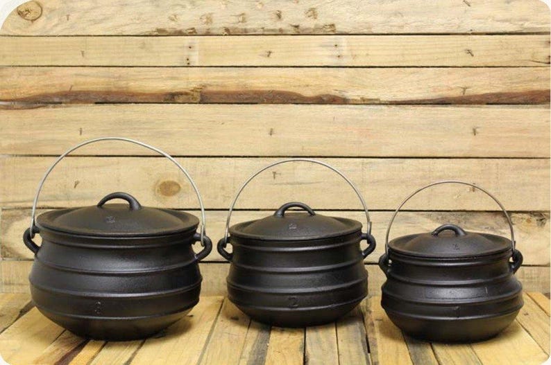 May include: Three black cast iron pots of varying sizes, each featuring a rounded body, a matching lid with a small handle, and a silver-coloured metal wire bail handle attached to two side lugs. The pots are arranged in a row.