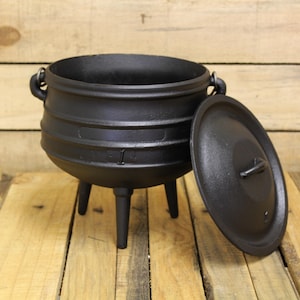 May include: A black cast iron cauldron with a lid, resting on three legs. The cauldron has two handles and a lid with a small handle. The cauldron and lid are on a wooden surface.