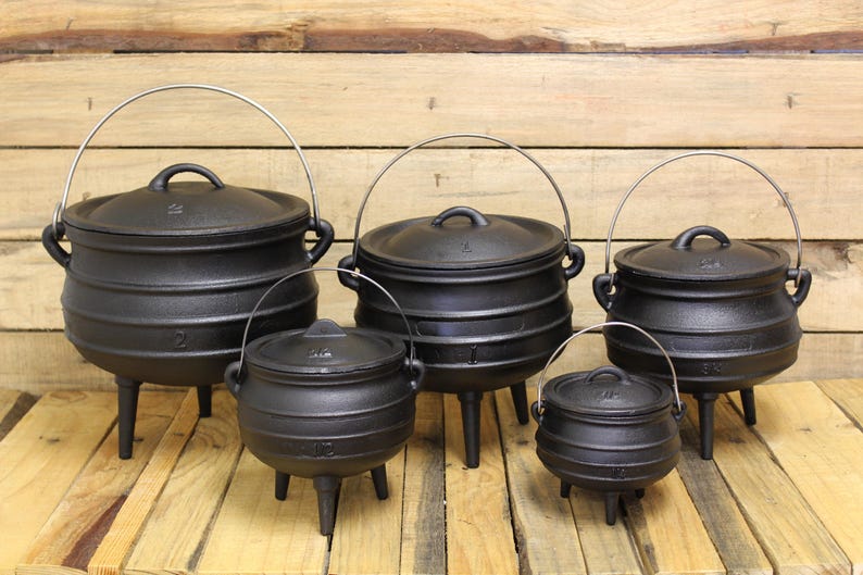 May include: A set of five black cast iron pots with lids, each featuring three legs and a metal wire handle. The pots vary in size and are arranged in two rows, displaying a rounded body shape with horizontal ridges on the exterior surface.