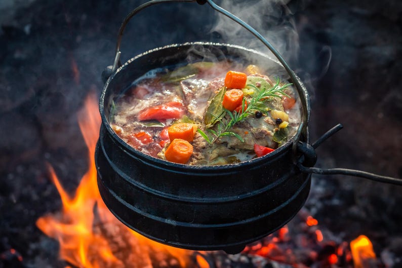 May include: A black cast iron pot with a metal handle suspended over an open fire. The pot contains a stew with visible orange carrot slices, red tomato pieces, green herbs, and dark peppercorns, with steam rising from the hot liquid.