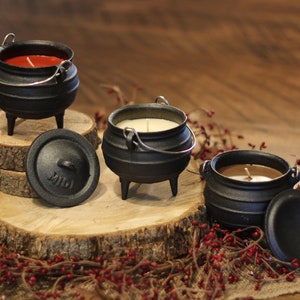 May include: Three black cast iron cauldron candles with lids. The candles are red, white, and brown. The candles are sitting on a wooden log slice.