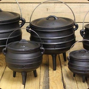 May include: A collection of black cast iron pots with lids, each featuring three legs and a wire handle. The pots have a rounded belly shape and are embossed with size markings such as 1/2 and 1, indicating their capacity in litres.
