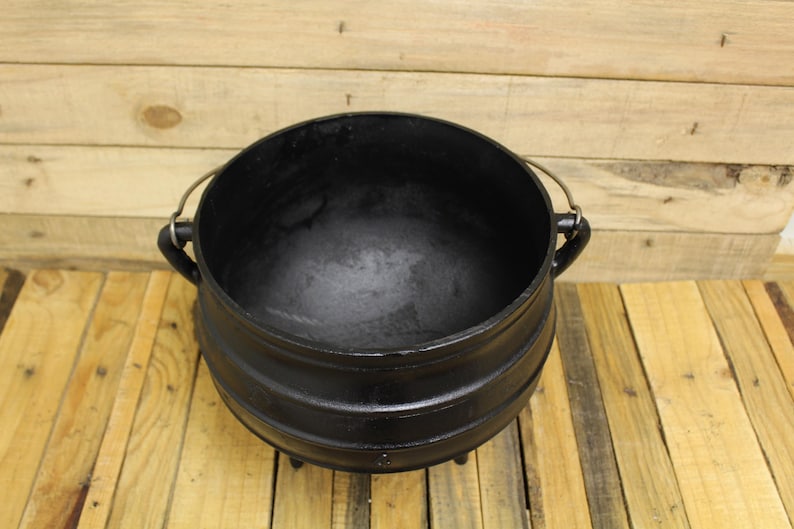 May include: A black cast iron pot with a rounded belly, a wide open top, and three short legs. The pot features two side handles and a thin, arched metal wire handle attached to the top, providing a sturdy structure for cooking or display.